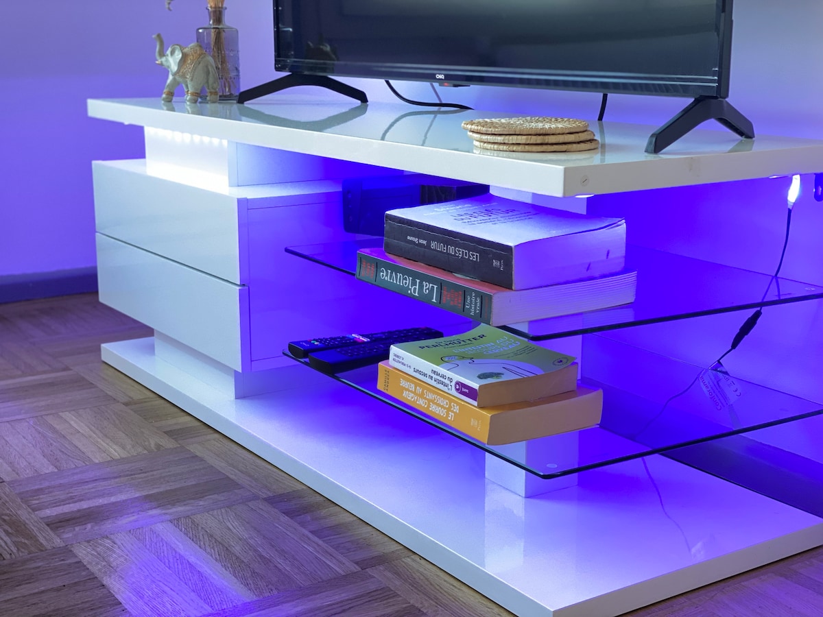A modern entertainment unit is displayed, featuring a sleek white finish. Beneath a television, glass shelves are illuminated in soft blue light, holding several books and a remote control. A decorative item is placed on top, adding to the unit's contemporary design.