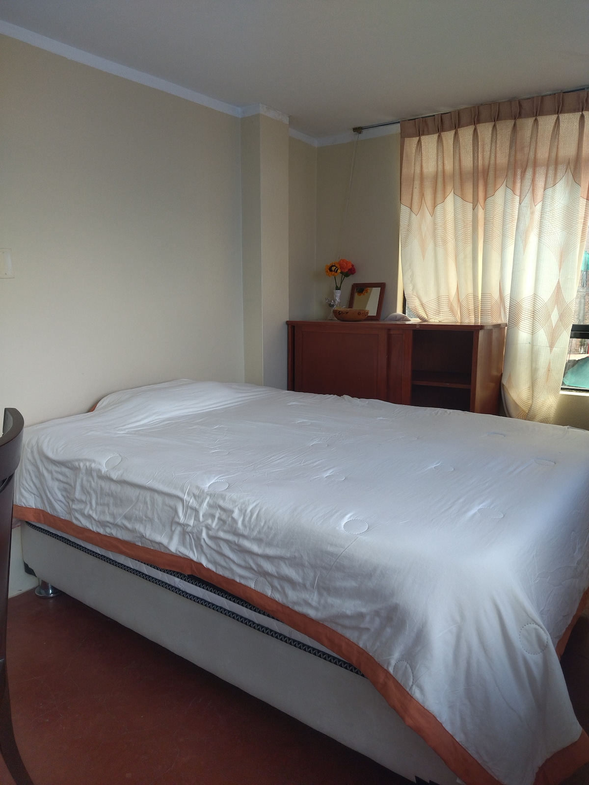 A cozy sleeping area features a spacious bed with a light-colored coverlet. A wooden cabinet is positioned nearby, while a window with sheer curtains allows natural light to enter, creating a calm and inviting atmosphere.