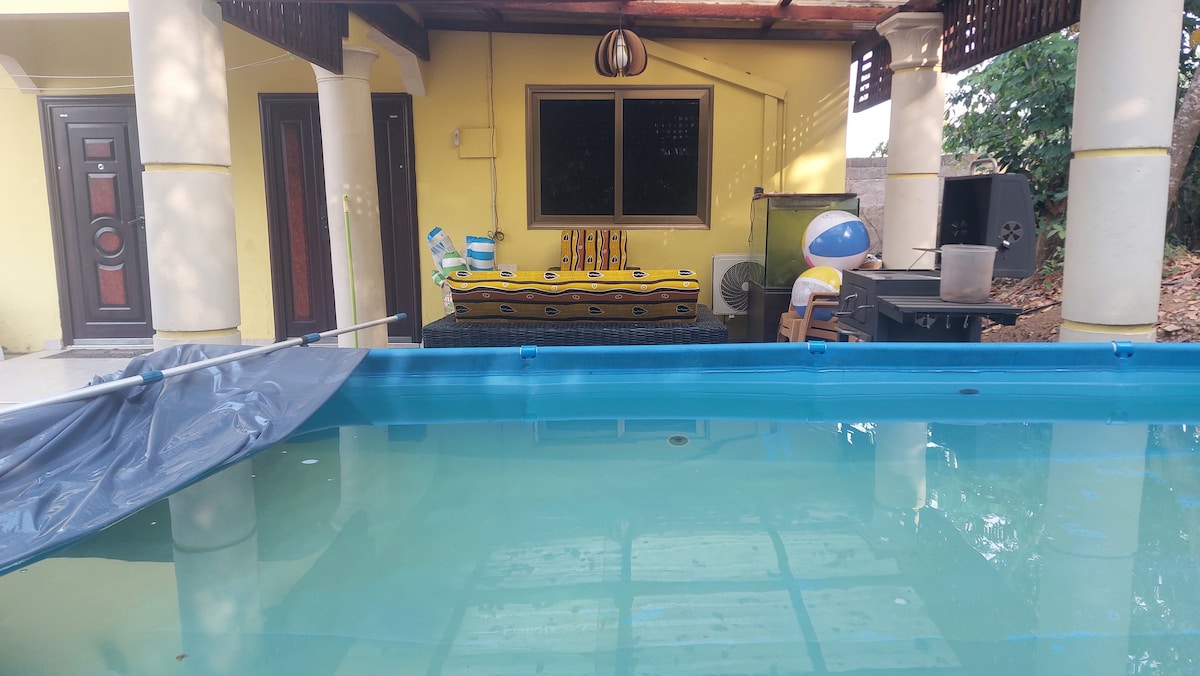 A swimming pool occupies the foreground, with a yellow inflatable boat visible on its edge. A patio area is seen in the background, featuring a grill and outdoor seating beneath a wooden awning, surrounded by greenery.