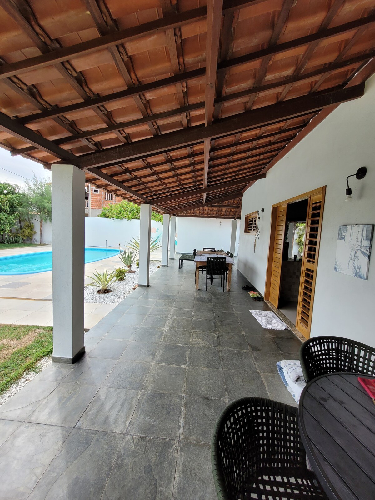 An outdoor patio is featured with a tiled floor and a large wooden table surrounded by black woven chairs. A covered area leads to the pool, with soft lighting illuminating the walls and plants nearby.