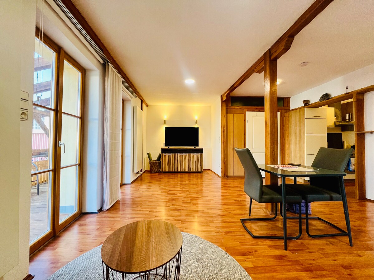 An open studio space displays a wooden floor, with natural light filtering through glass doors. A dining area is set with a table and chairs, while a cozy seating area includes a TV mounted on a wooden console. The overall ambiance suggests comfort and functionality.