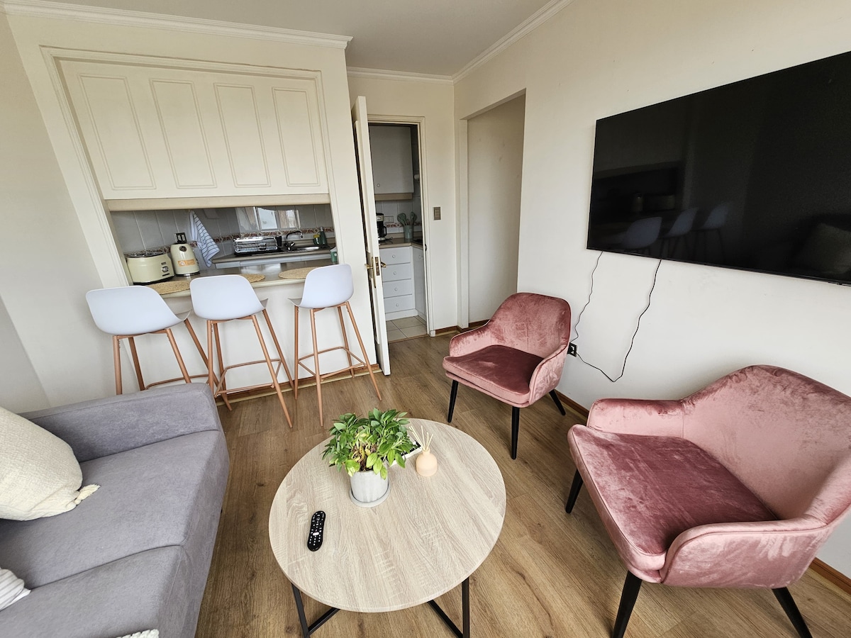 A cozy living area features a grey sofa and two pink upholstered chairs surrounding a round wooden coffee table. A flat-screen television is mounted on the wall, and a compact kitchen area with high stools is visible in the background, enhancing the open layout.
