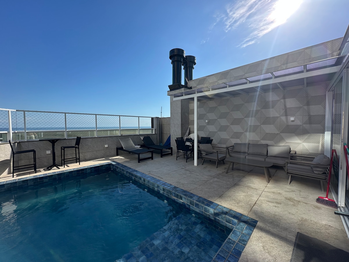 A spacious terrace features a private pool surrounded by lounge chairs and a shaded seating area. The geometric patterned wall provides a modern backdrop, while the coastline can be seen in the distance under a clear blue sky.