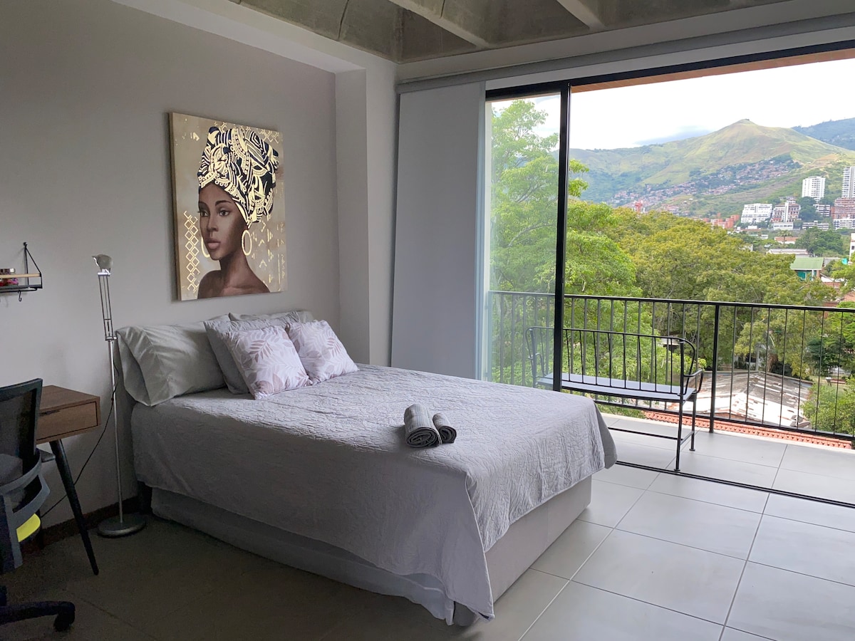 A bright room features a spacious bed dressed in light linens, complemented by decorative pillows. A large window offers a view of nearby mountains and greenery. A compact desk and chair are positioned nearby, enhancing functionality while natural light floods the space.