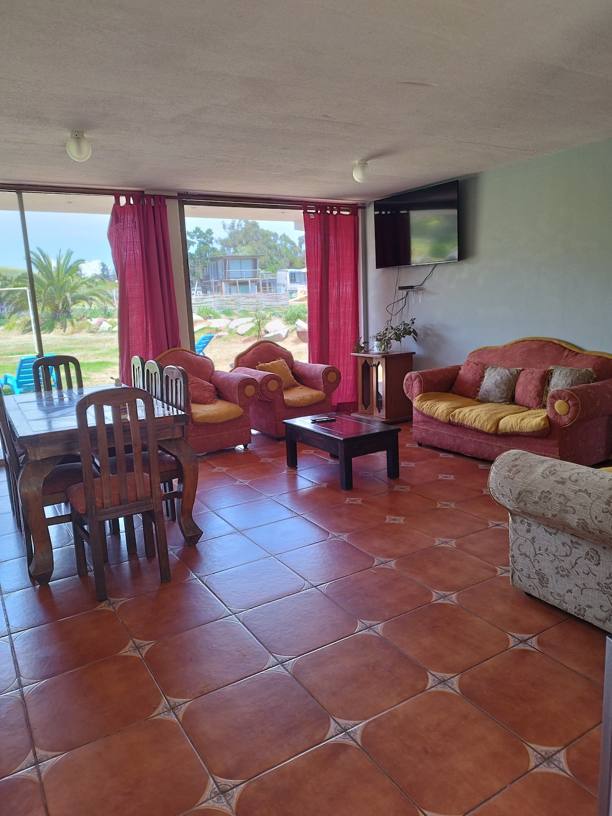 A welcoming living area features a dining table surrounded by wooden chairs. Two sofas, upholstered in rich fabric with yellow cushions, complement a coffee table at the center. Large windows allow natural light to fill the space, with outdoor views visible through stylish red curtains.