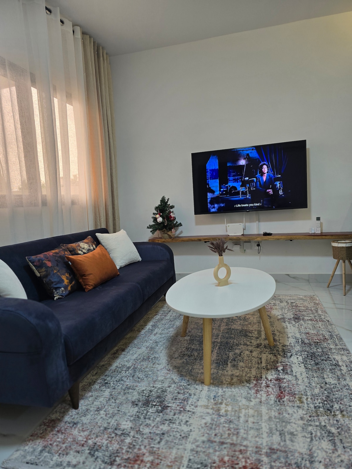 A cozy living area is displayed, featuring a navy blue sofa adorned with decorative pillows in warm tones. A round coffee table sits in front, placed on a textured area rug. A wall-mounted television is visible, accompanied by a small Christmas tree.