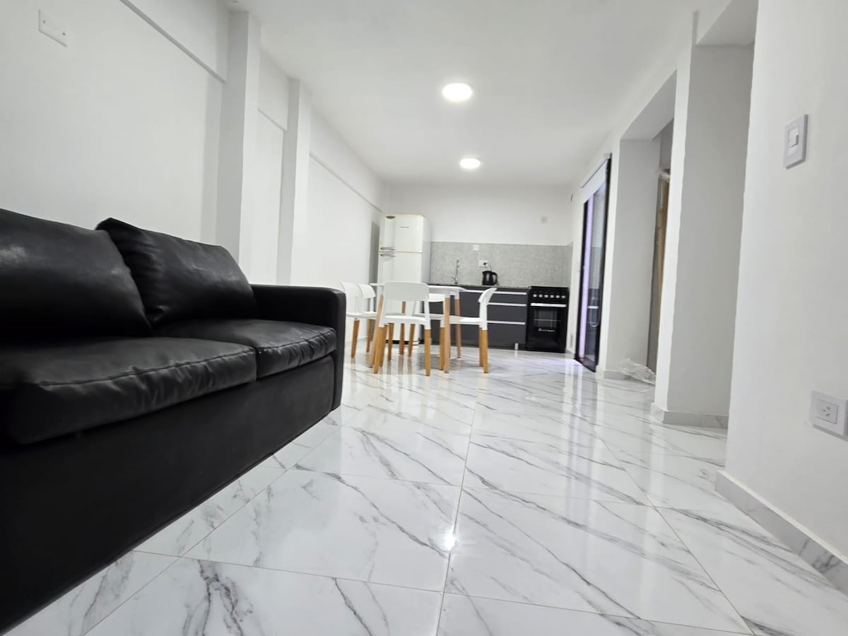 An open-plan living area features a sleek black sofa positioned in the foreground. A dining table with white chairs is situated in the center, and a modern kitchen area is visible in the background, showcasing light gray cabinetry and appliances against a tiled backsplash.