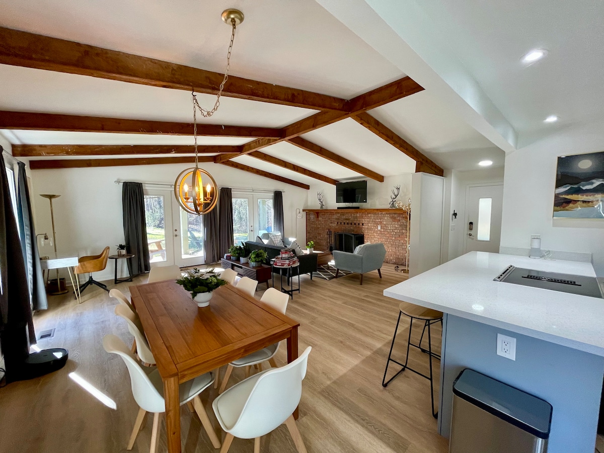 An open living and dining area showcases exposed wooden beams and large windows, allowing natural light to fill the space. A dining table with white chairs and a modern kitchen island are visible, along with a cozy seating arrangement around a brick fireplace.