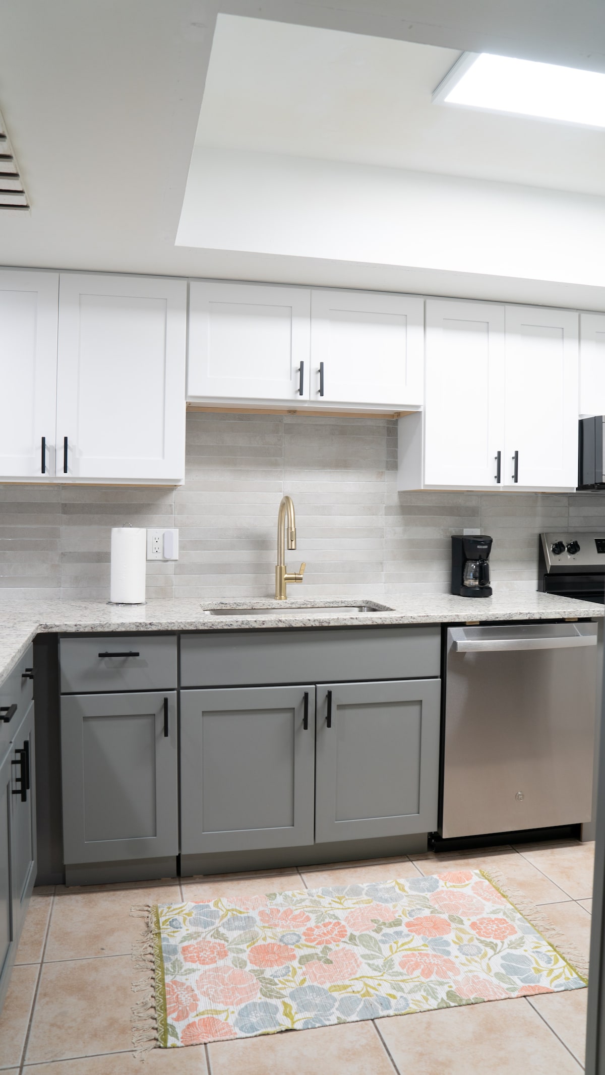 A modern kitchen features a blend of white and gray cabinetry, complemented by a sleek granite countertop. A gold faucet adds a touch of elegance. Appliances include a coffee maker and a silver dishwasher. A floral-patterned rug adds a pop of color to the tiled floor.
