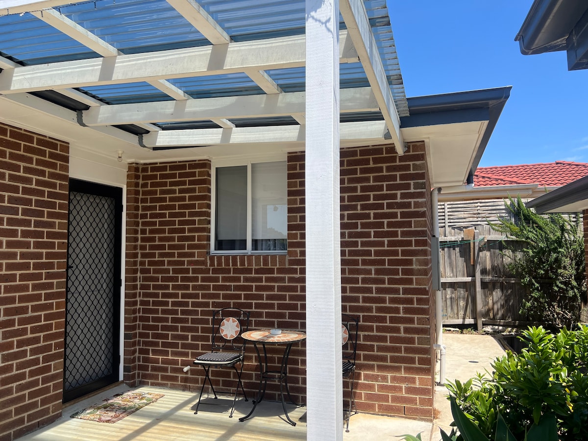 An outdoor patio area features a small table and two chairs, ideal for relaxation. The space is shaded by a translucent roof, with brick walls adding warmth. Nearby, greenery and a wooden fence provide a sense of privacy, complemented by clear blue skies above.