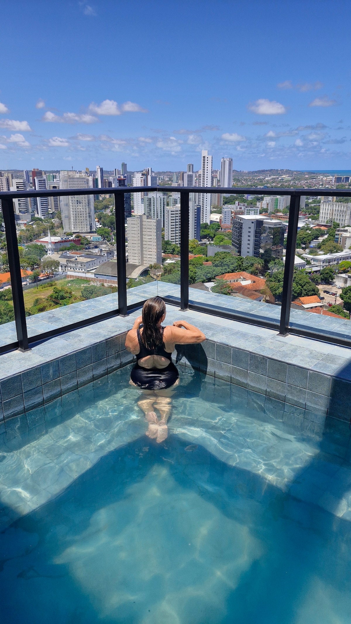 A rooftop pool is depicted with a person enjoying the view while seated at the edge. The water appears clear, reflecting the vibrant blue sky. A panoramic cityscape, featuring tall buildings and greenery, is visible in the background.