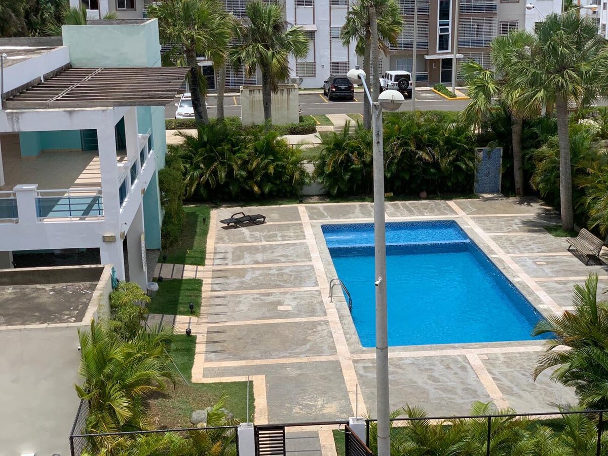 A clear view of a swimming pool is surrounded by well-maintained greenery and palm trees. Spacious deck areas with lounge chairs are positioned near the pool. The surrounding structures display modern architecture and provide a sense of community living.