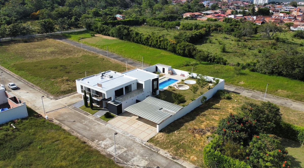 An aerial view of a modern house set in a spacious, green landscape. The property includes a private pool and jacuzzi, surrounded by neatly landscaped gardens. Surrounding areas of grass and some distant urban structures can be seen, highlighting the serene environment.