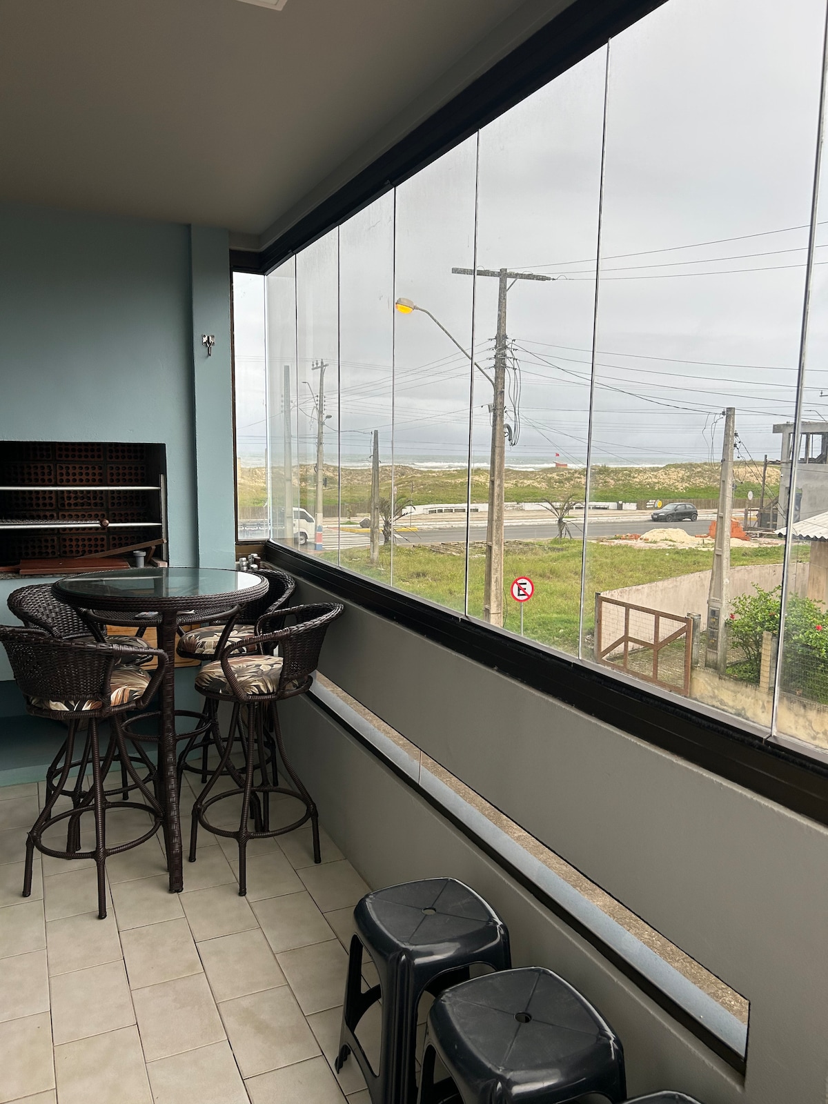 A balcony area features a round table surrounded by high-backed chairs. Large glass panels allow for an unobstructed view of the outside scenery, which includes a road and greenery. The floor is tiled, and additional seating is present in the form of stools.