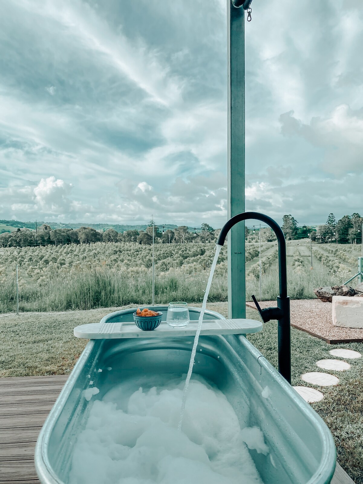 A freestanding bathtub is set on a wooden deck, with bubbles visible and water flowing from a sleek black faucet. A bowl of snacks and a drink sit on the edge. In the background, a view of lush fields and distant hills is framed by a cloudy sky.