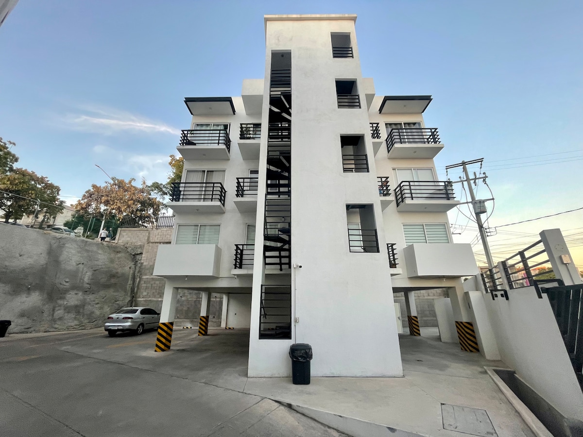 The exterior of a modern three-story building is depicted, featuring a clean white facade and several balconies. Stairs are visible on one side, leading to the upper levels. Parking spaces are located below the building, with a single vehicle parked in the designated area.