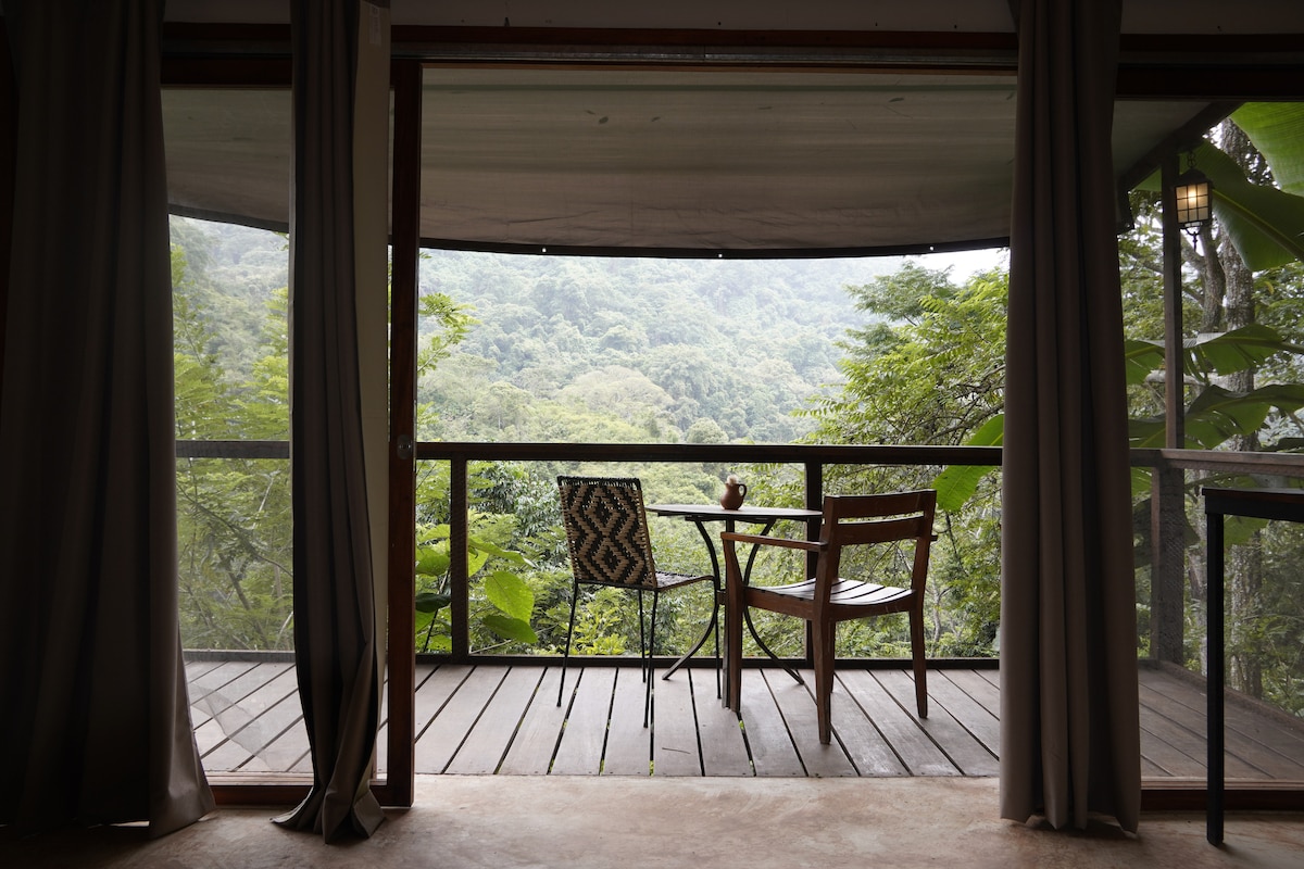 A balcony is featured, providing a view of the surrounding mountains and lush greenery. Two wooden chairs and a small table are positioned on the balcony, framed by floor-to-ceiling windows and soft, heavy drapes.