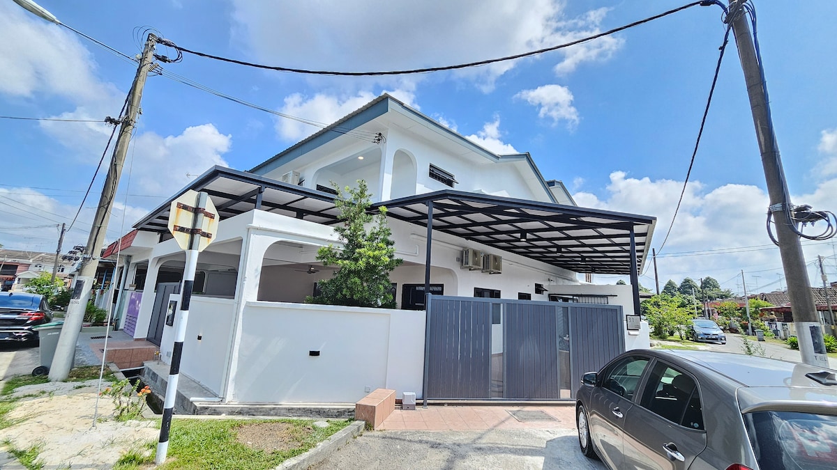 The exterior of the townhouse is shown, featuring a covered parking area and a landscaped front garden. The structure has a modern design with white walls and large windows, while surrounding spaces indicate a vibrant urban setting.