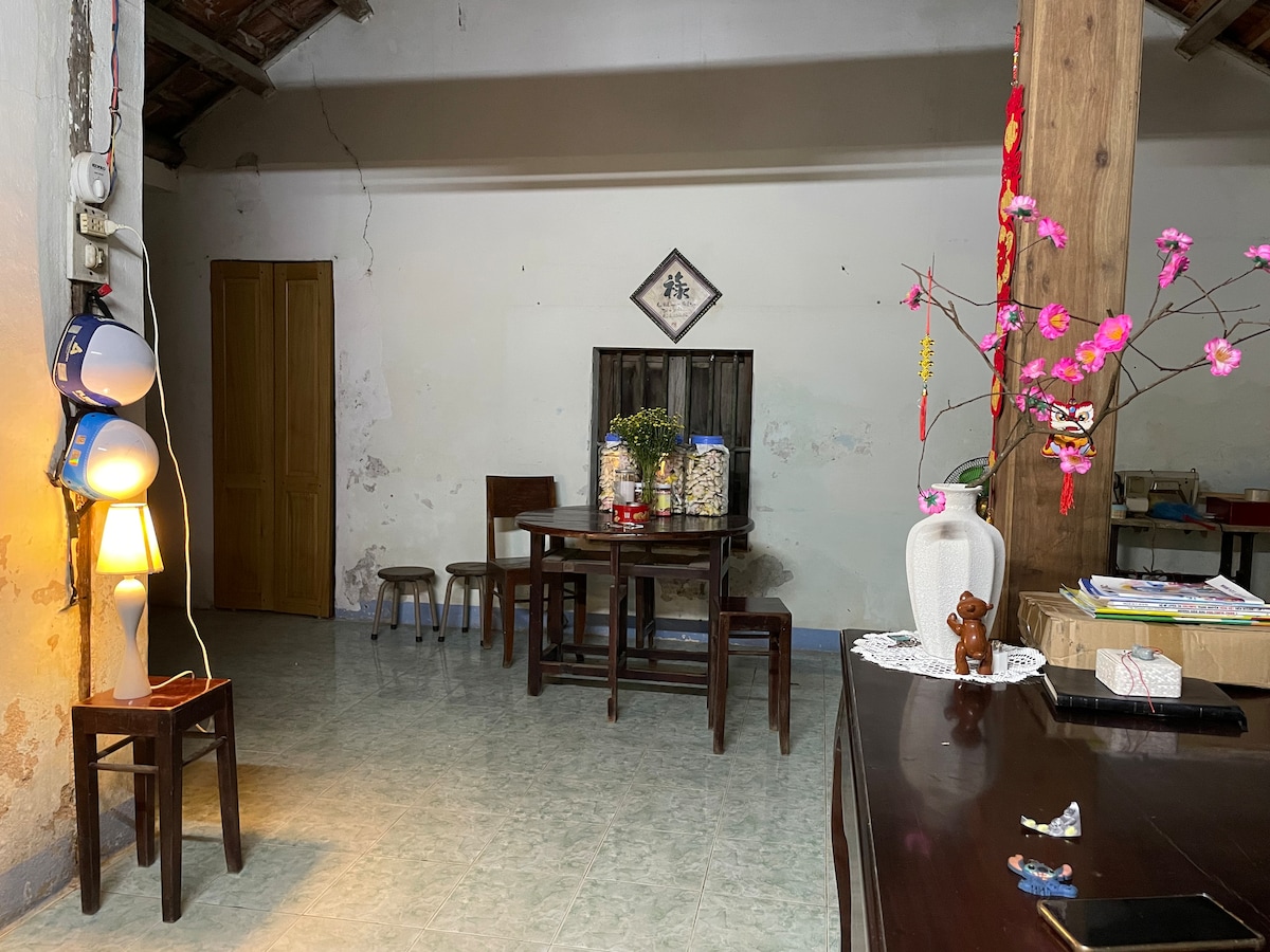 A dining area is furnished with a wooden table surrounded by four chairs. A small decorative tree with pink blossoms is positioned nearby. A lamp stands on the left, providing ambient lighting. The walls exhibit a rustic finish, complementing the wooden elements.