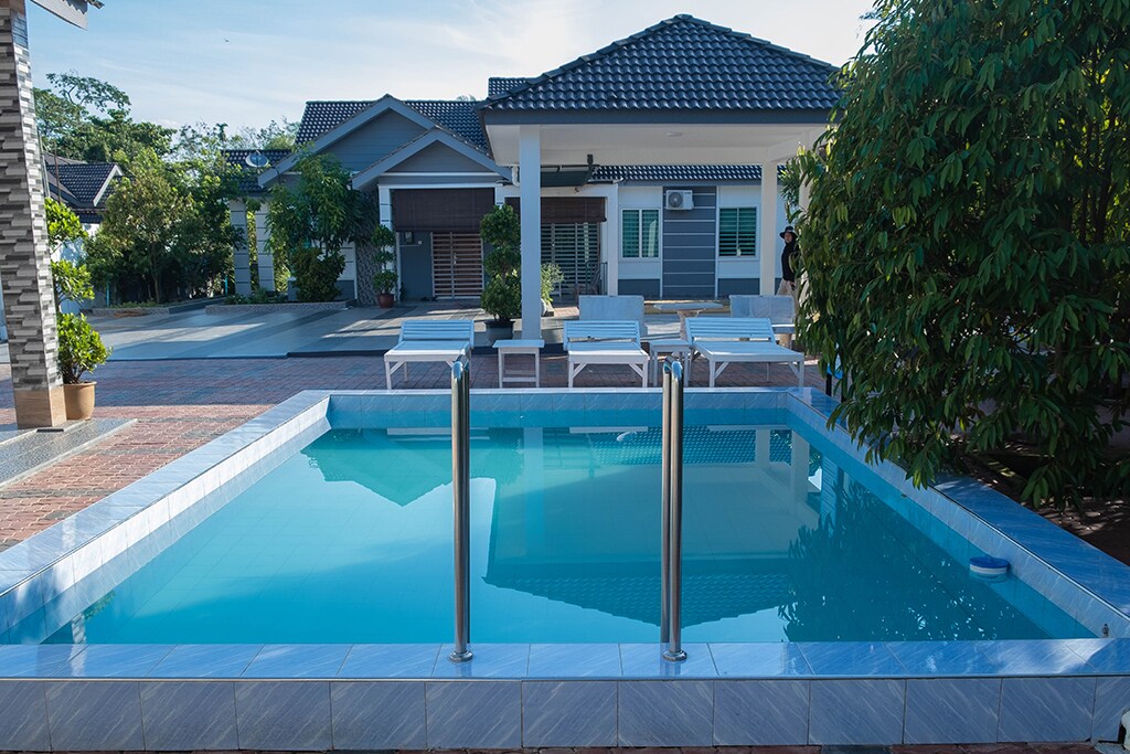 A serene pool area is featured, surrounded by a sunlit patio. Two ladders provide access to the clear blue water. Several lounge chairs are positioned nearby, creating a relaxing outdoor space. A comfortable residential building is visible in the background.