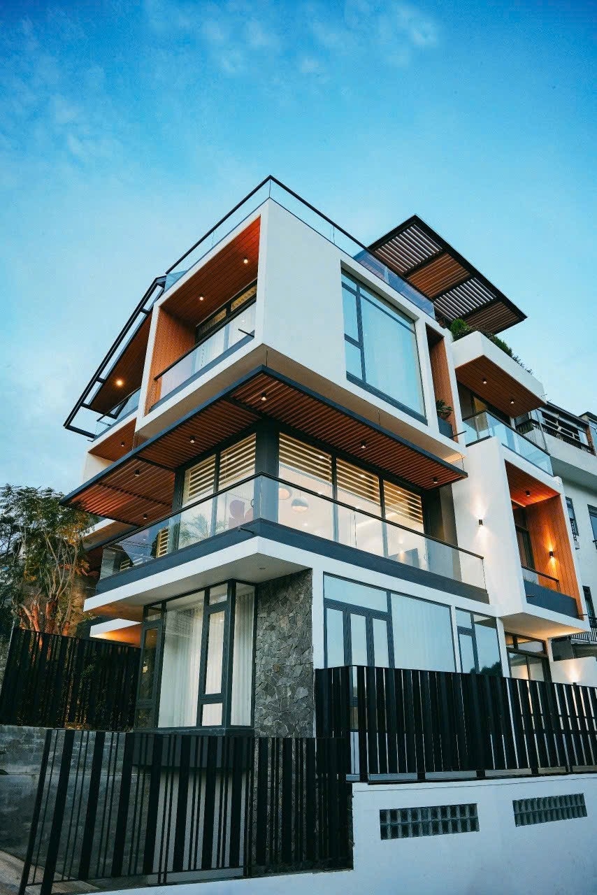 A modern villa is showcased from an exterior angle, featuring multiple levels with large windows and a blend of materials. The structure is framed by a black fence, with landscaped elements visible in the background, against a clear blue sky.