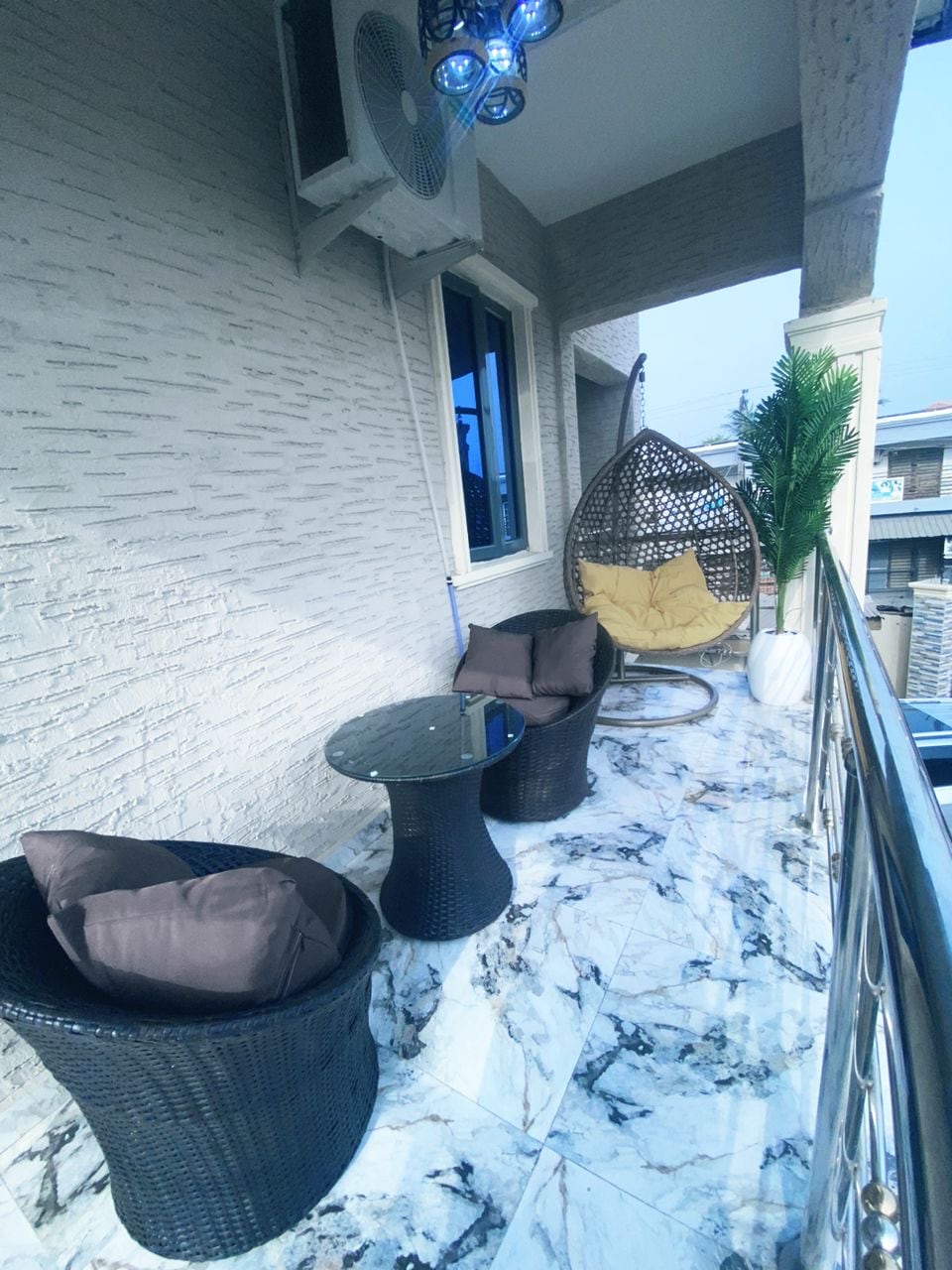 A balcony space features a circular hanging chair alongside two woven armchairs. A round glass table rests on a marbled floor, while decorative green foliage adds a touch of nature. Sliding glass doors lead to the interior, allowing for natural light.
