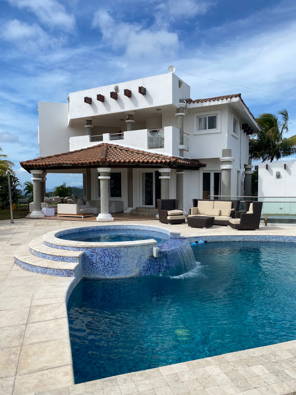 A multi-level home is depicted, featuring a large swimming pool and a jacuzzi surrounded by a patio area. Elegant outdoor furniture is arranged for relaxation. The exterior showcases a mix of white walls and a terracotta roof, set against a clear blue sky and green palm trees.
