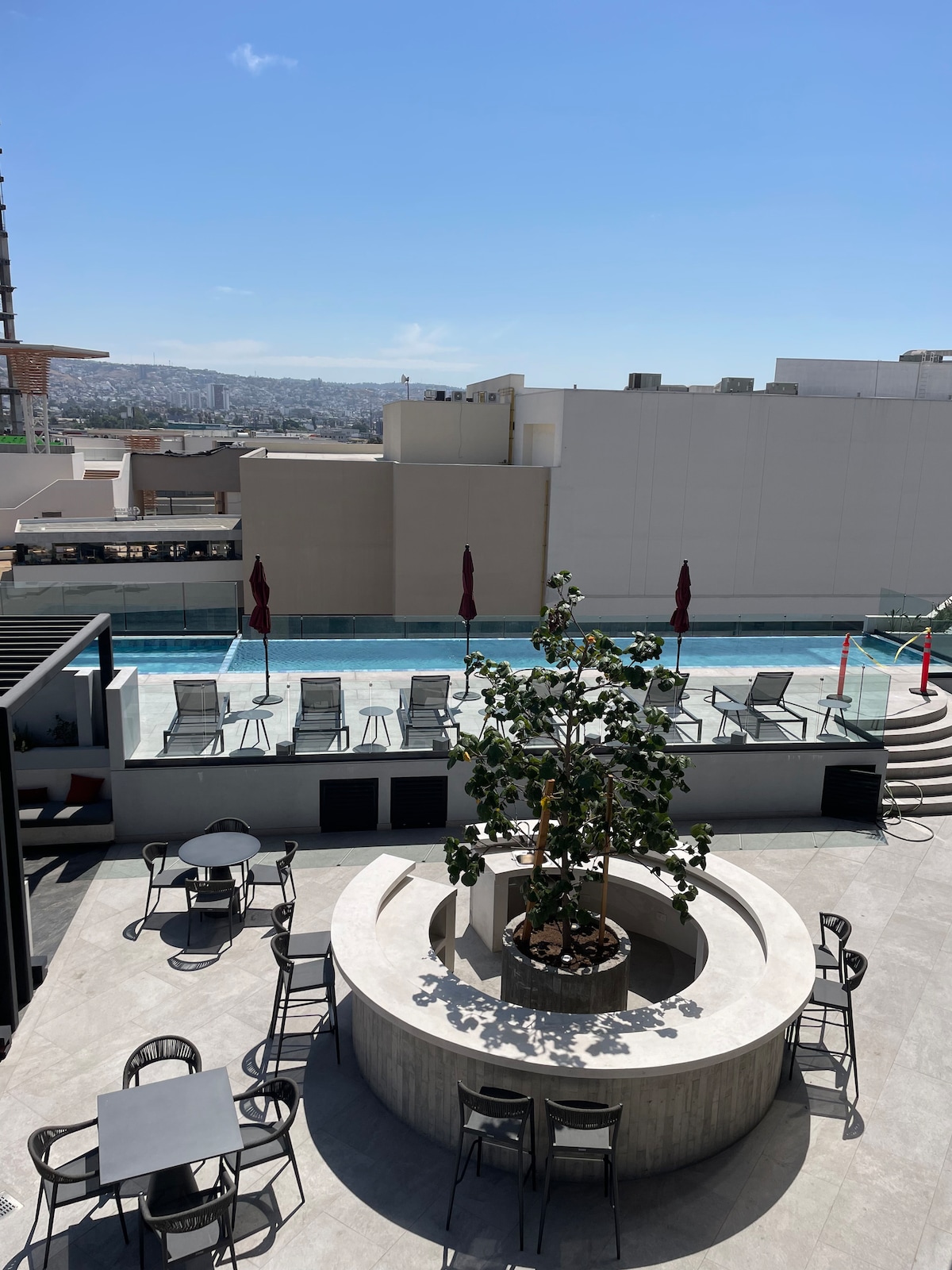 An outdoor terrace showcases a central planter with a leafy tree, surrounded by various seating options. A serene pool is visible in the background, with lounge chairs arranged neatly. The clear blue sky enhances the ambiance, providing a spacious and inviting environment.