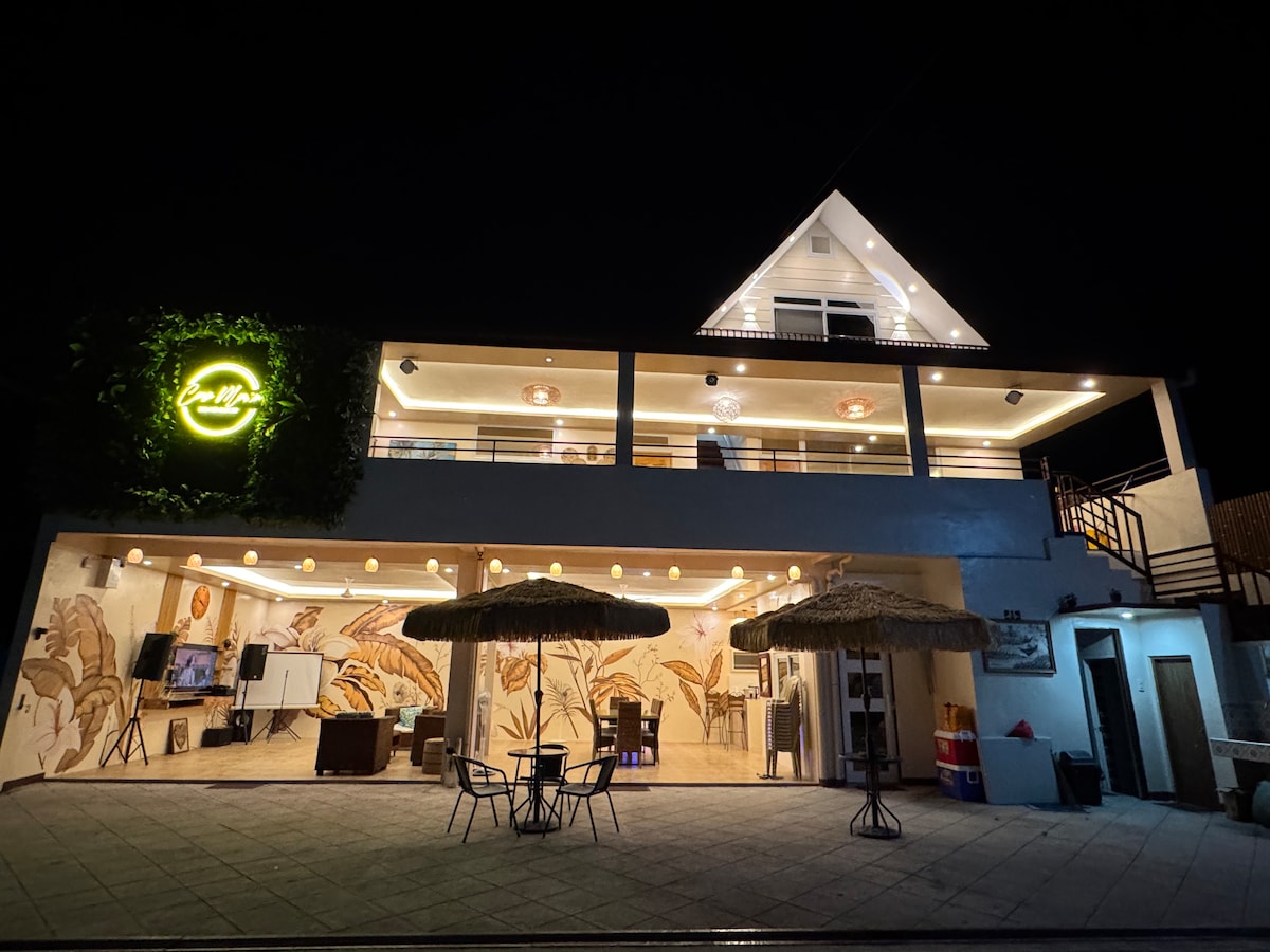 The property showcases a modern facade illuminated by a large neon sign. Outdoor seating is arranged under two thatched umbrellas, creating a relaxed atmosphere. The spacious patio features a mural and lighting that enhances the inviting space for social gatherings.