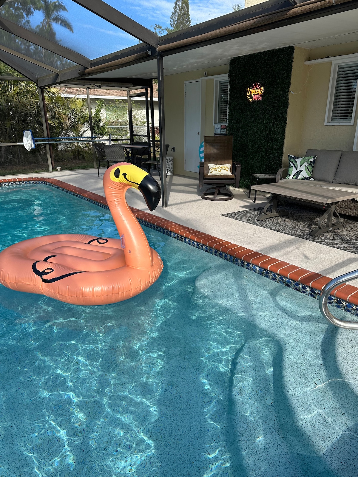 A large heated pool features a pink flamingo float resting on the clear blue water. Sunlight sparkles across the surface, while lounge chairs and a side table are positioned nearby under a screened enclosure, providing a relaxed setting for outdoor enjoyment.