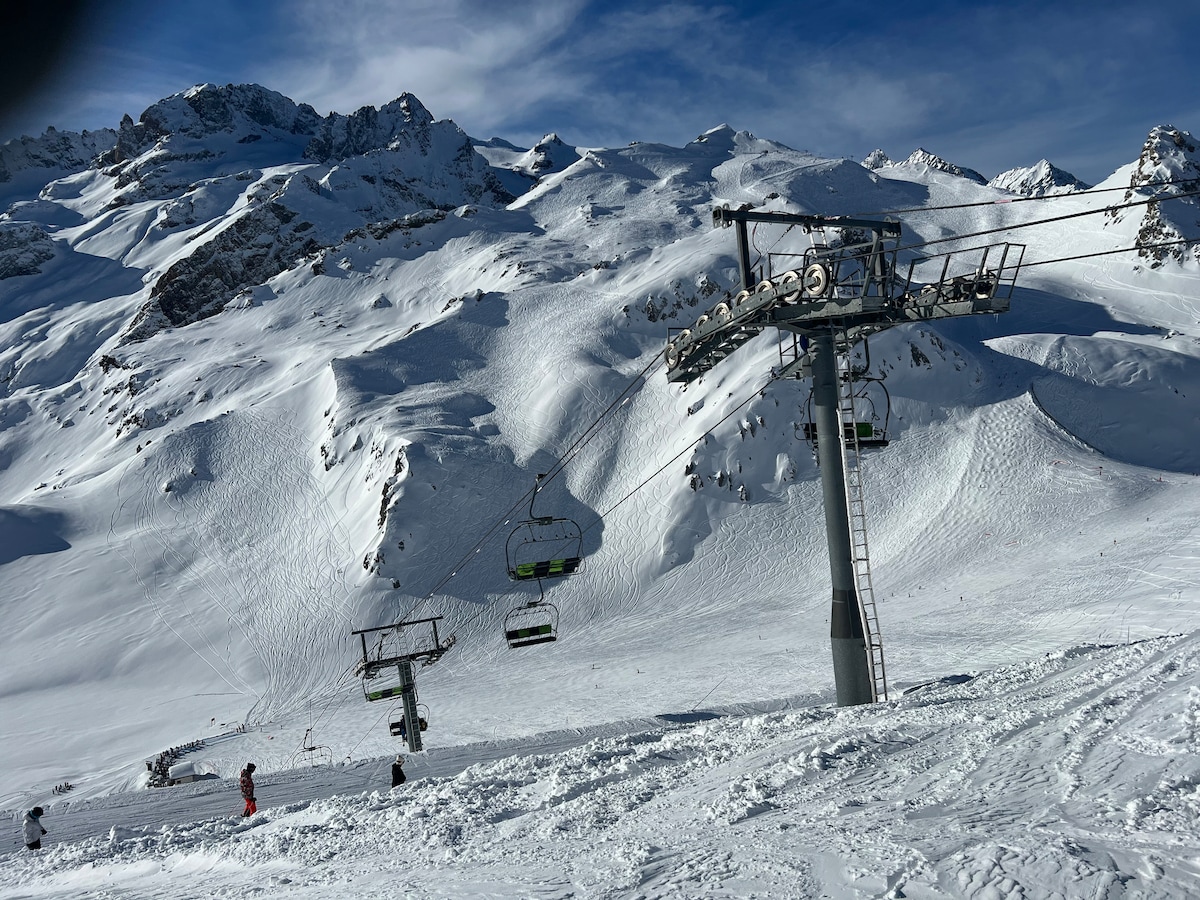 A ski lift is seen ascending a snowy mountain slope, surrounded by towering snow-capped peaks. The bright blue sky contrasts with the white landscape. Several skiers are positioned at the base of the lift, preparing to embark on their descent.