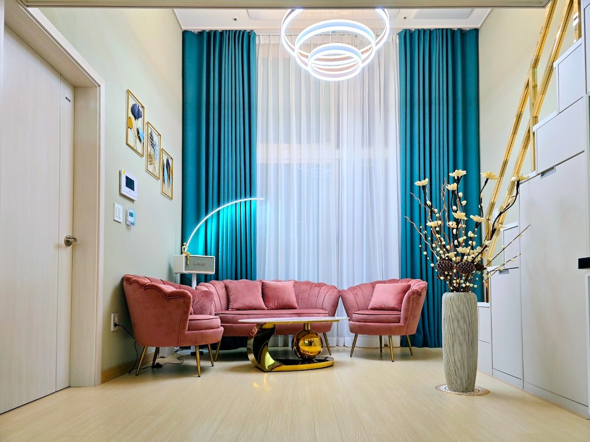 The living area presents a modern aesthetic with a pink curved sofa set, positioned around a stylish coffee table. Large windows are dressed with light curtains, allowing soft natural light to enter. A decorative vase with branches and a contemporary light fixture enhance the space.