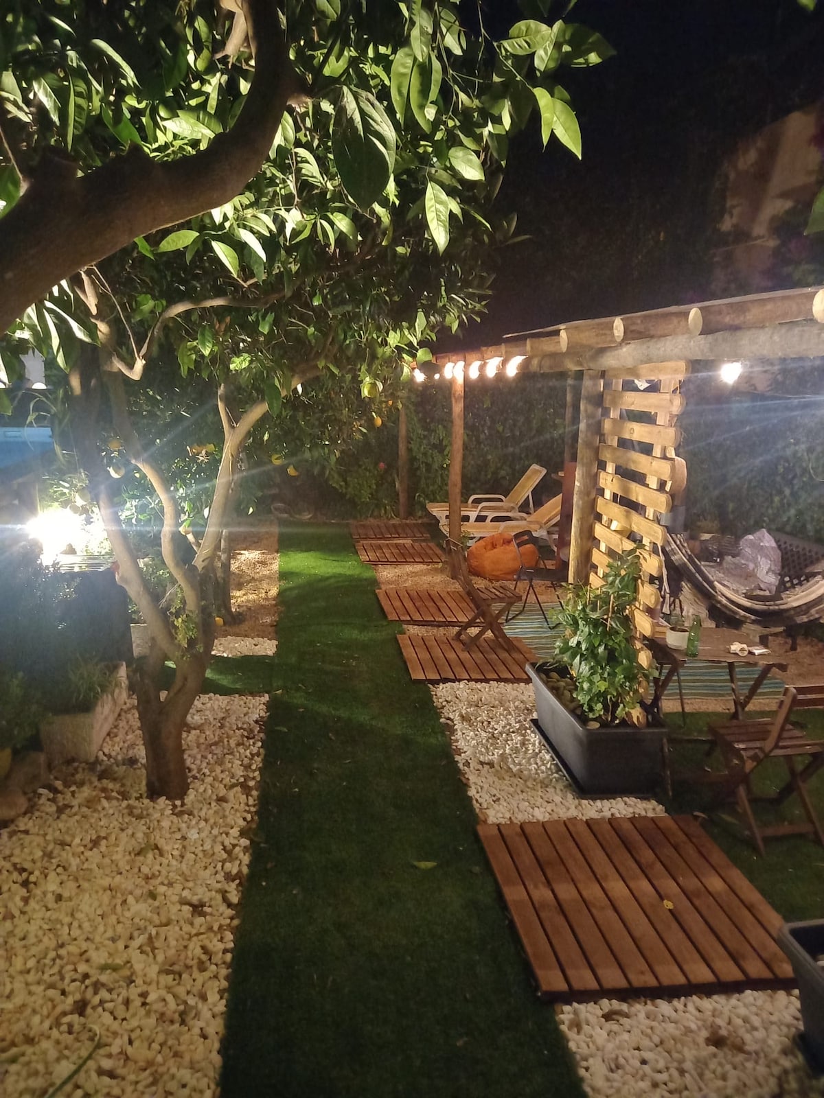 A landscaped garden is illuminated by soft lighting, featuring a pathway lined with wooden decking and stones. Comfortable seating options, including lounge chairs and a hammock, are present among the greenery, creating a serene outdoor space for relaxation.