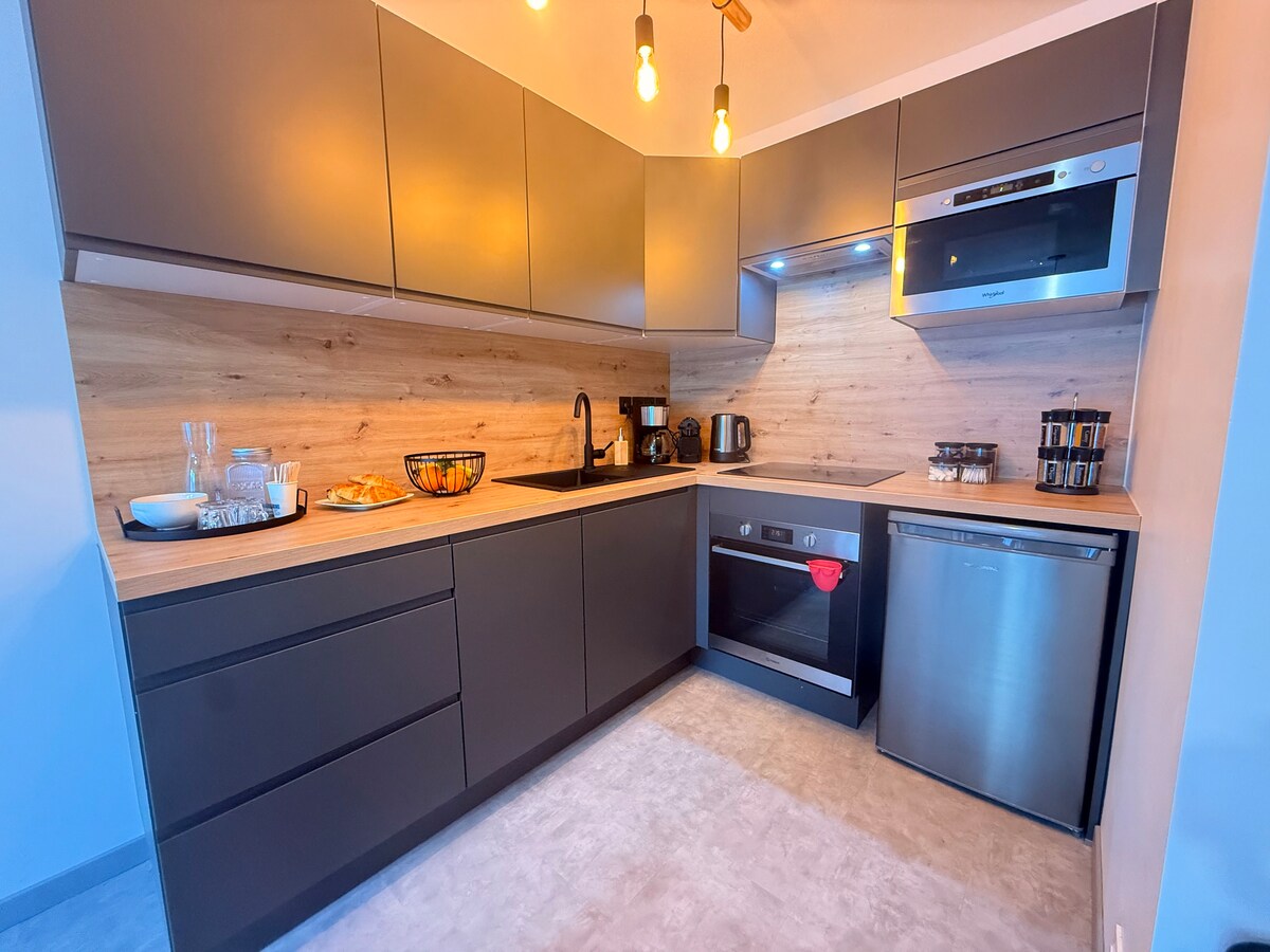 A modern kitchen is presented with sleek grey cabinetry and wooden accents. It features essential appliances including an oven, refrigerator, and coffee maker. The space is well-lit with pendant lighting, and a bowl of fresh fruit is positioned on the countertop, enhancing the functional design.
