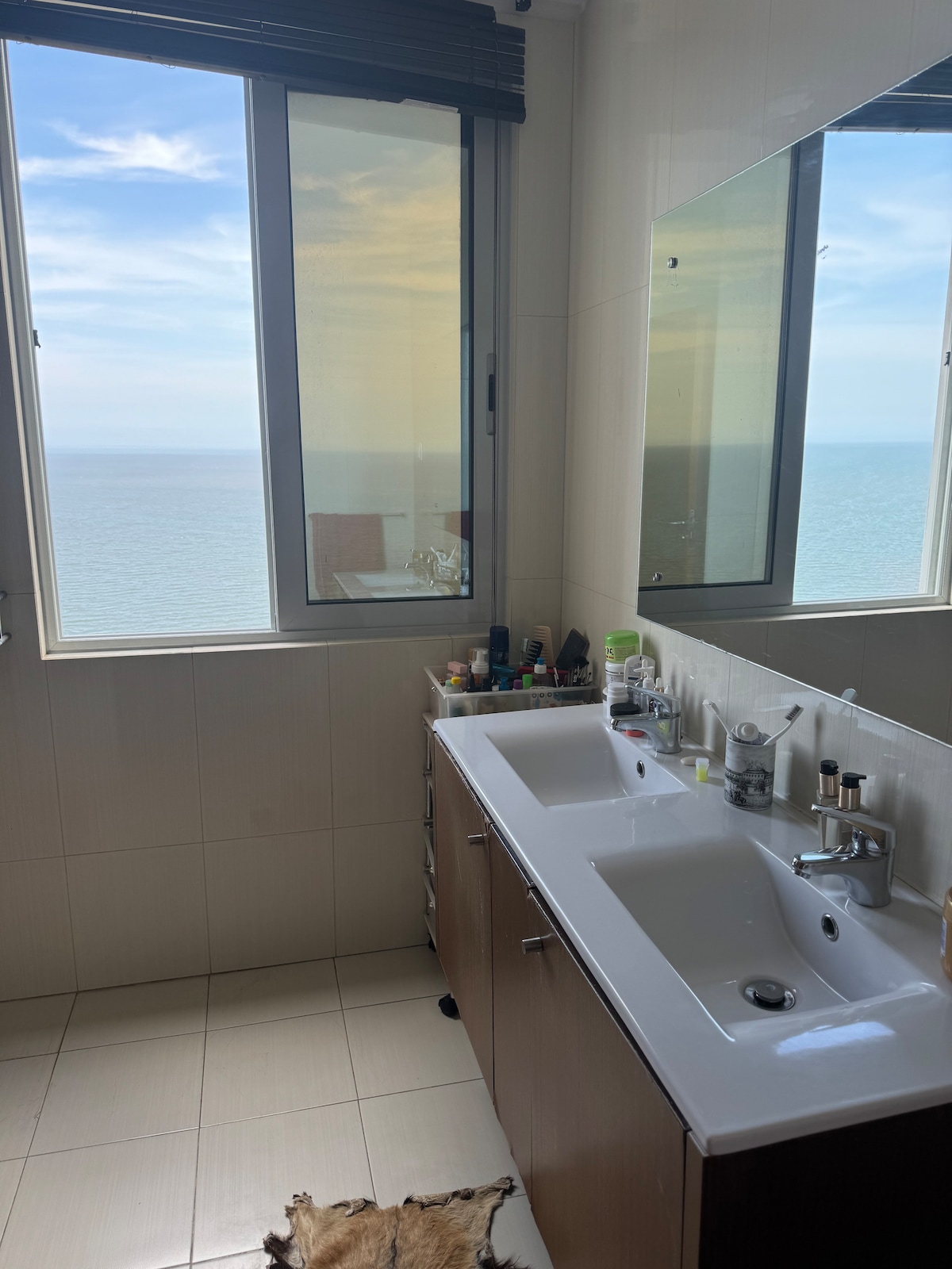 A well-lit bathroom features a double sink set against a sleek countertop. Large windows offer a view of the ocean, while natural light reflects off the glossy tiles. Personal items and toiletries are neatly arranged along the counter for easy access.