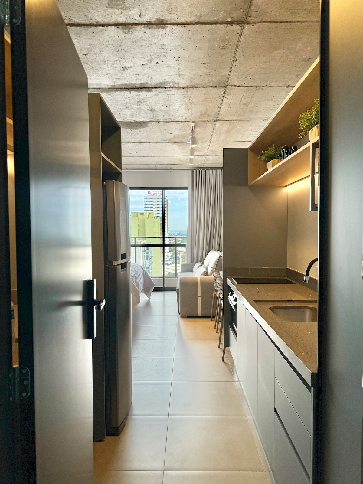 A view from the entrance reveals a stylish kitchenette with a refrigerator and counter space, leading to a cozy seating area. Natural light enters through large windows, illuminating the room and highlighting urban scenery in the background.
