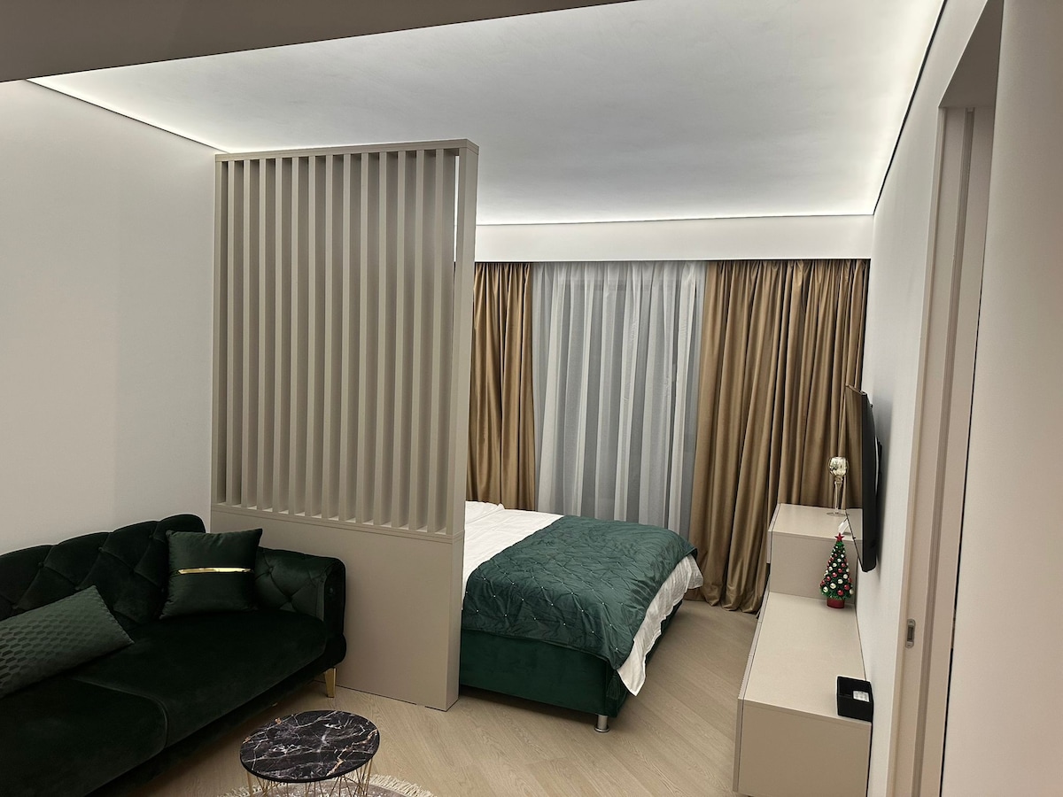 A studio layout features a green velvet sofa complemented by decorative cushions. A spacious bed with a textured green cover is situated behind a partially open room divider. Light-colored walls and soft drapes create a serene atmosphere, while a simple bedside table completes the setting.