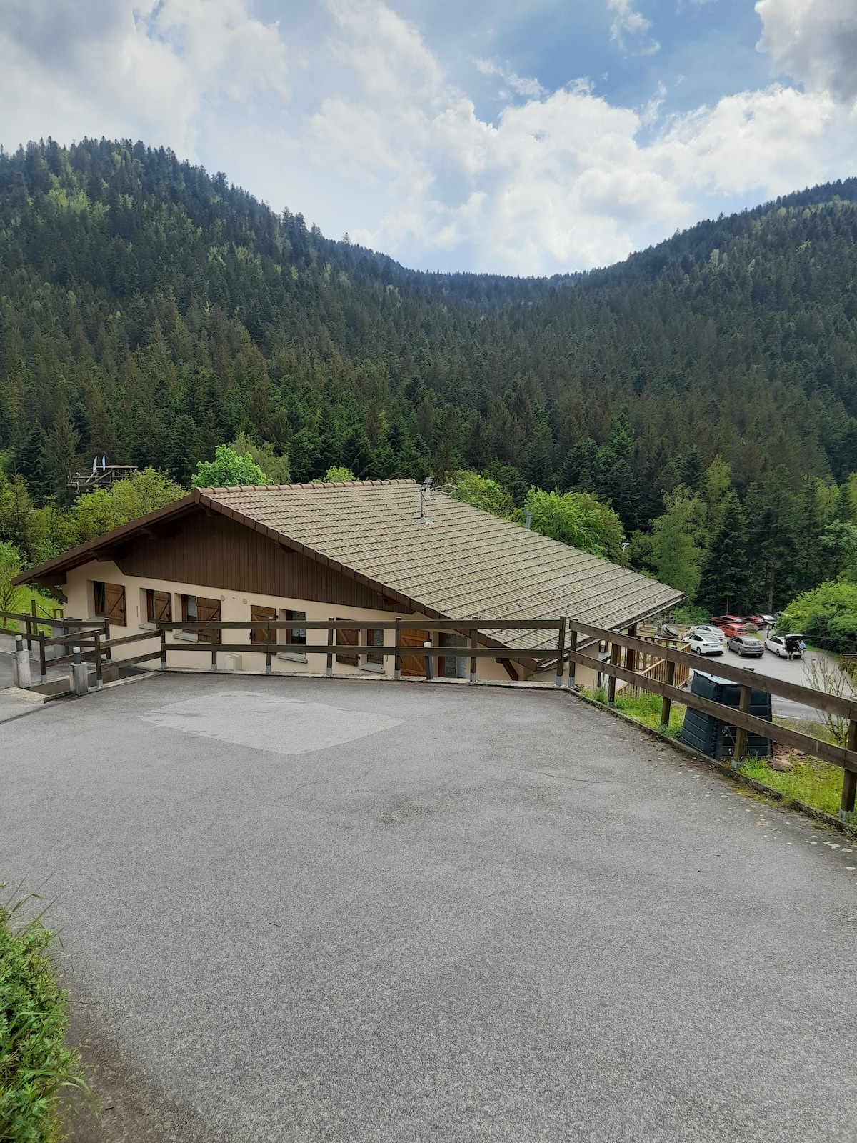A chalet-style building rests against a backdrop of dense green mountains. The structure features a sloped roof and multiple wooden-paneled windows. A gravel path leads to the entrance, with wooden railings lining the sides. Cars are visible parked nearby among the trees.