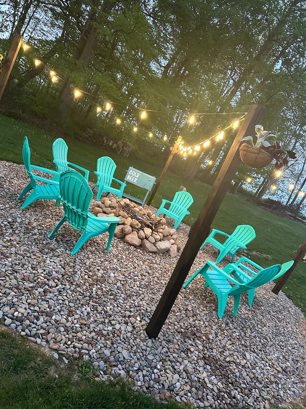 An outdoor seating area is surrounded by a gravel base, featuring multiple turquoise chairs arranged in a circular layout around a stone fire pit. String lights hang above, illuminating the space, while greenery and trees frame the background.