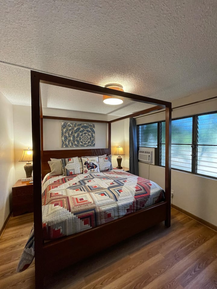 Chambre principale paisible avec un lit king size confortable, un éclairage doux et des alizés rafraîchissants à travers les grandes fenêtres à persiennes — parfait pour les nuits reposantes de Maui.
