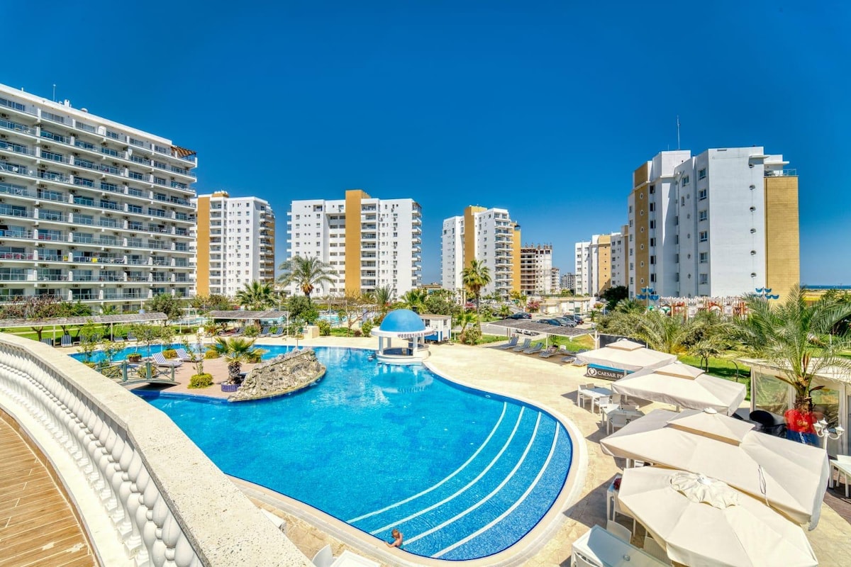 A spacious outdoor pool is surrounded by lounging areas and umbrellas, complemented by palm trees and landscaped greenery. Towering residential blocks provide a backdrop, with clear blue skies above enhancing the vibrant atmosphere of the Caesar Resort.