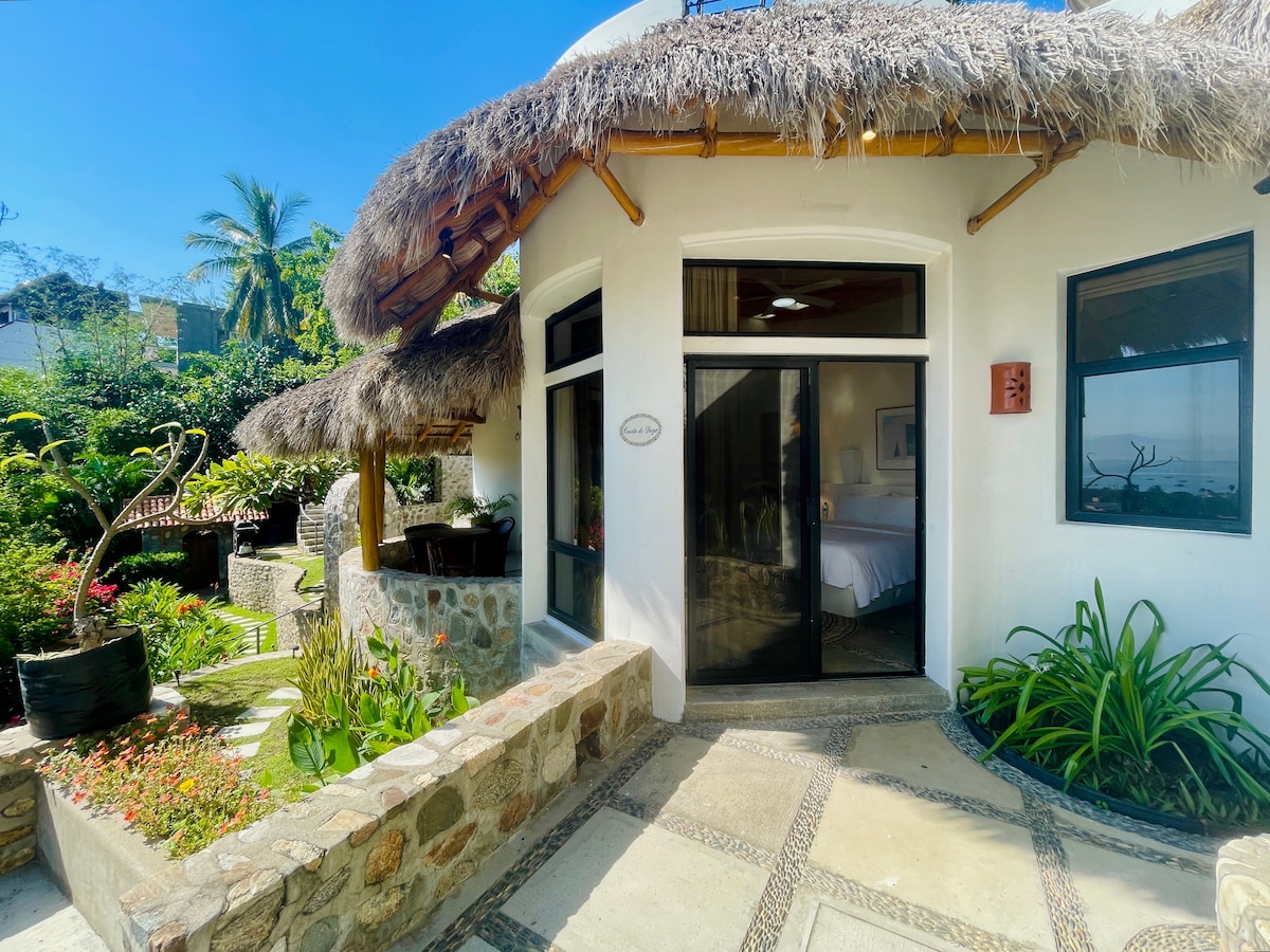 An exterior view of the villa displays a thatched roof structure surrounded by lush greenery and colorful flowers. The entrance is framed by stone pathways, leading to a glass door that provides a glimpse of the interior space and a spacious king bed.