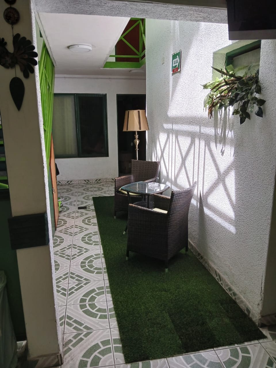 An intimate seating area is presented, featuring two wicker chairs and a glass table on a green carpet. Natural light filters through a nearby window, creating a bright but relaxed ambiance. The walls are adorned with greenery, offering a refreshing touch to the space.
