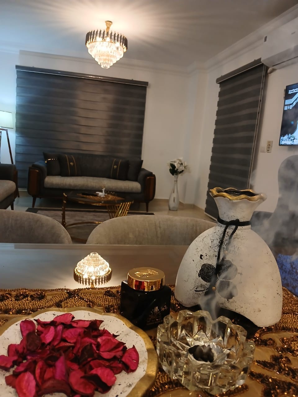 A living area is visible, featuring a stylish sofa and elegant lighting from a chandelier overhead. A table is adorned with a decorative bowl filled with dried rose petals and a lit incense holder, creating a calming ambiance. The backdrop includes soft gray curtains.