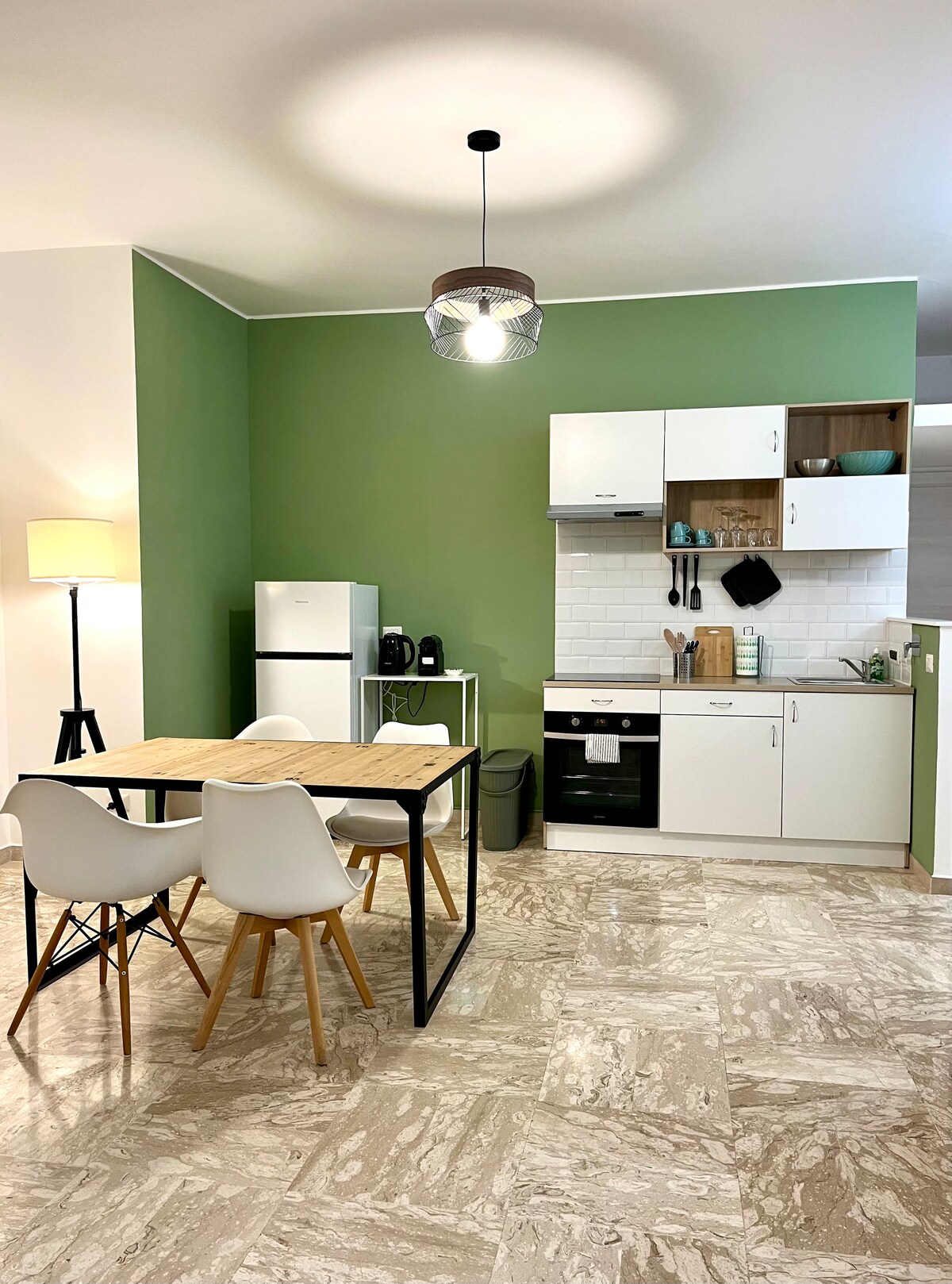 A modern kitchen area features green accent walls and a compact dining table with four chairs. Appliances, including a refrigerator and coffee maker, are visible. A light fixture hangs above the table, illuminating the space, while natural light enters from a nearby window.