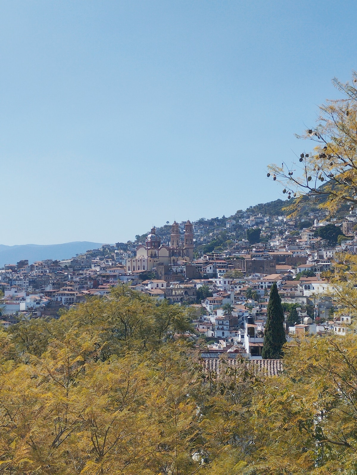 A panoramic view of a hillside town is presented, showcasing a variety of buildings nestled among lush greenery. The prominent Church of Santa Prisca stands out against the backdrop of a clear blue sky, framed by golden foliage.