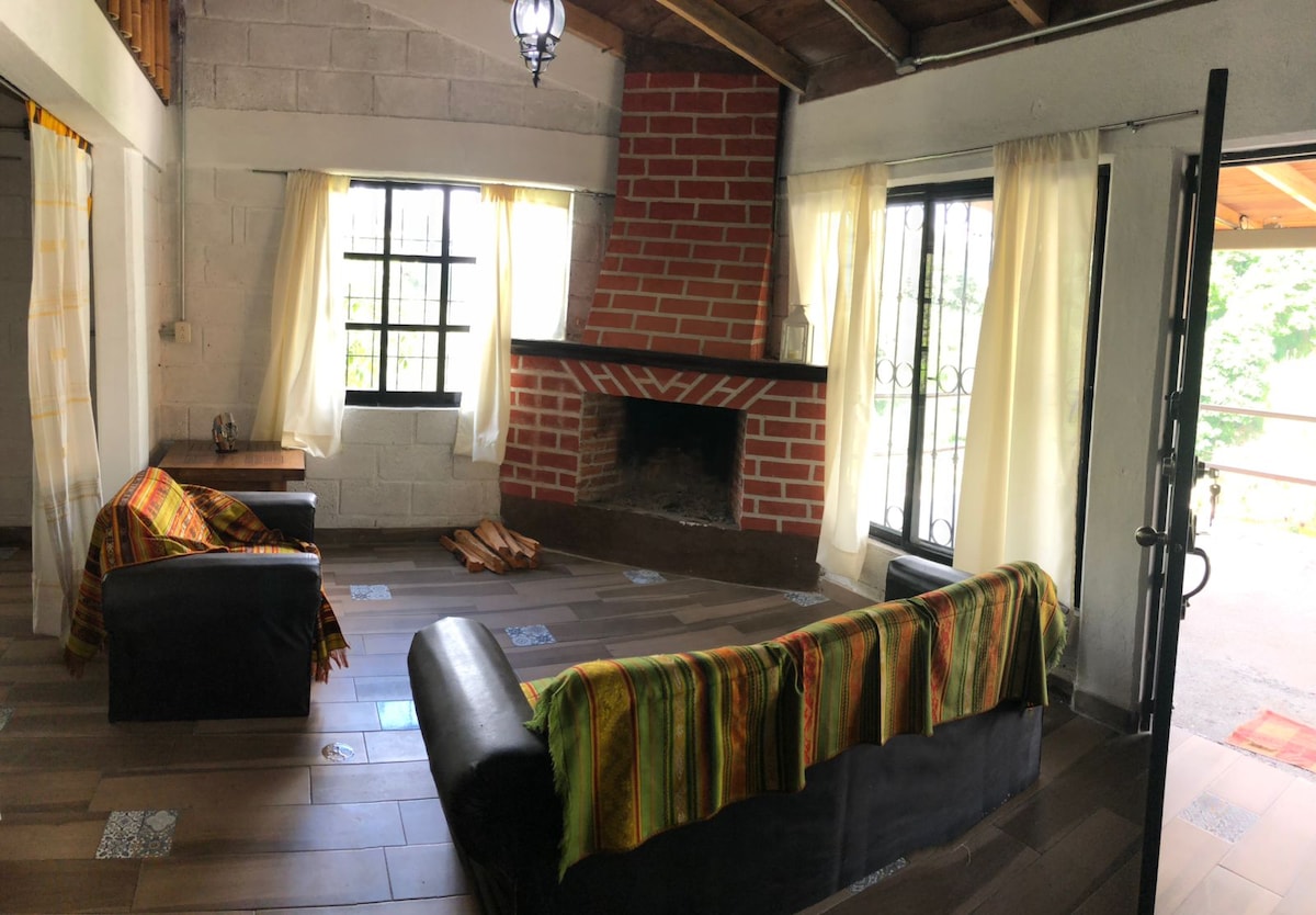 A warm living space is highlighted by a brick fireplace and large windows allowing natural light to fill the room. Two dark sofas are adorned with colorful woven throws. The rustic wooden ceiling and tiled floor contribute to a cozy atmosphere.