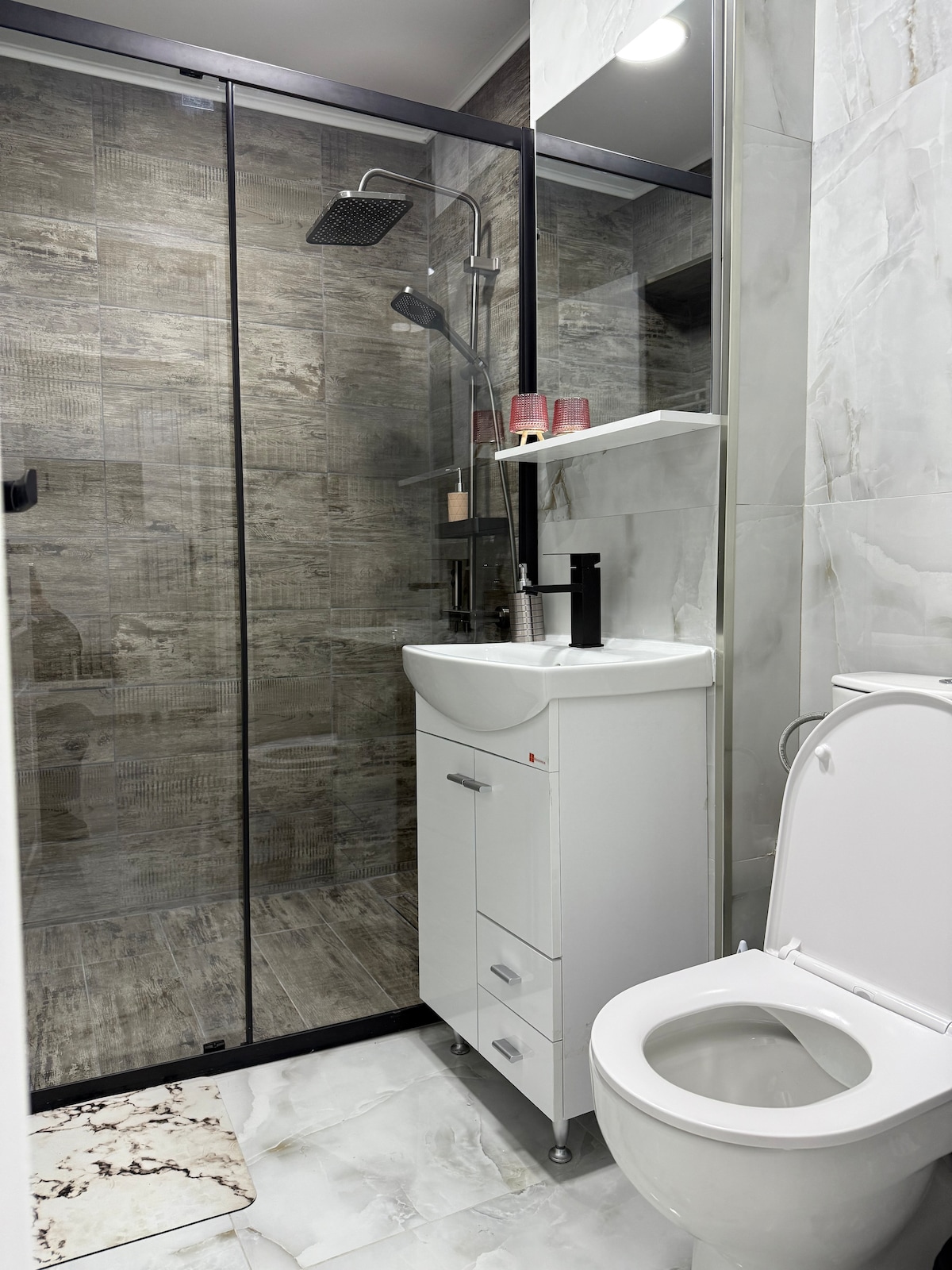 A modern bathroom is equipped with a glass-enclosed shower, featuring a rainfall showerhead. A compact white vanity with drawers provides storage, and a toilet is positioned nearby. Neutral tiles in various shades adorn the walls and floor, creating a clean and functional space.
