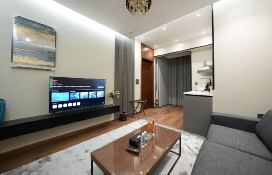 A modern living area features a sleek gray couch facing a wall-mounted television. Soft lighting illuminates the space, highlighting a glass coffee table placed on a light-colored area rug. A small dining table can be seen in the background near the kitchen area.