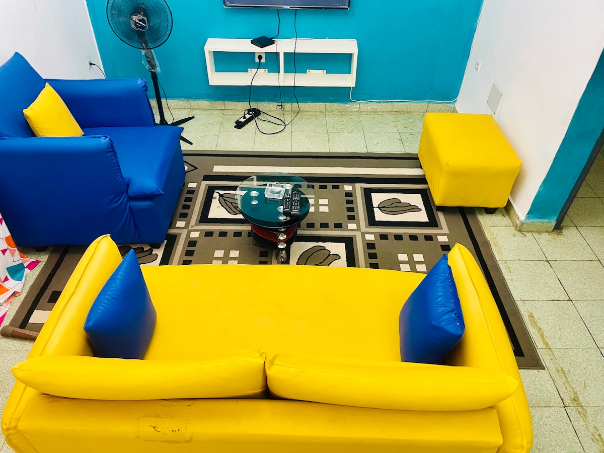 A vibrant living area is highlighted by a yellow sofa with blue accent pillows and matching armchairs. A rectangular coffee table rests in the center, with a patterned rug providing visual interest on the floor. A wall-mounted TV and a fan are visible, contributing to the space's functionality.